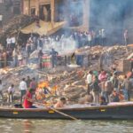 Varanasi, India - December 21, 2019: A cremation  scene, where people are burnt near Ganges River at Manikarnika Ghat, the ashes are taken by the wind, people sing while they bring the dead ones to the cremation, a boat passes near ghat and all is covered in smoke, in the holy city of Varanasi, India.