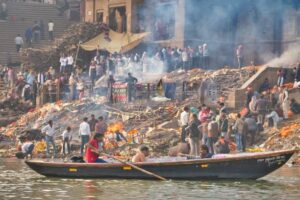 Varanasi, India - December 21, 2019: A cremation  scene, where people are burnt near Ganges River at Manikarnika Ghat, the ashes are taken by the wind, people sing while they bring the dead ones to the cremation, a boat passes near ghat and all is covered in smoke, in the holy city of Varanasi, India.